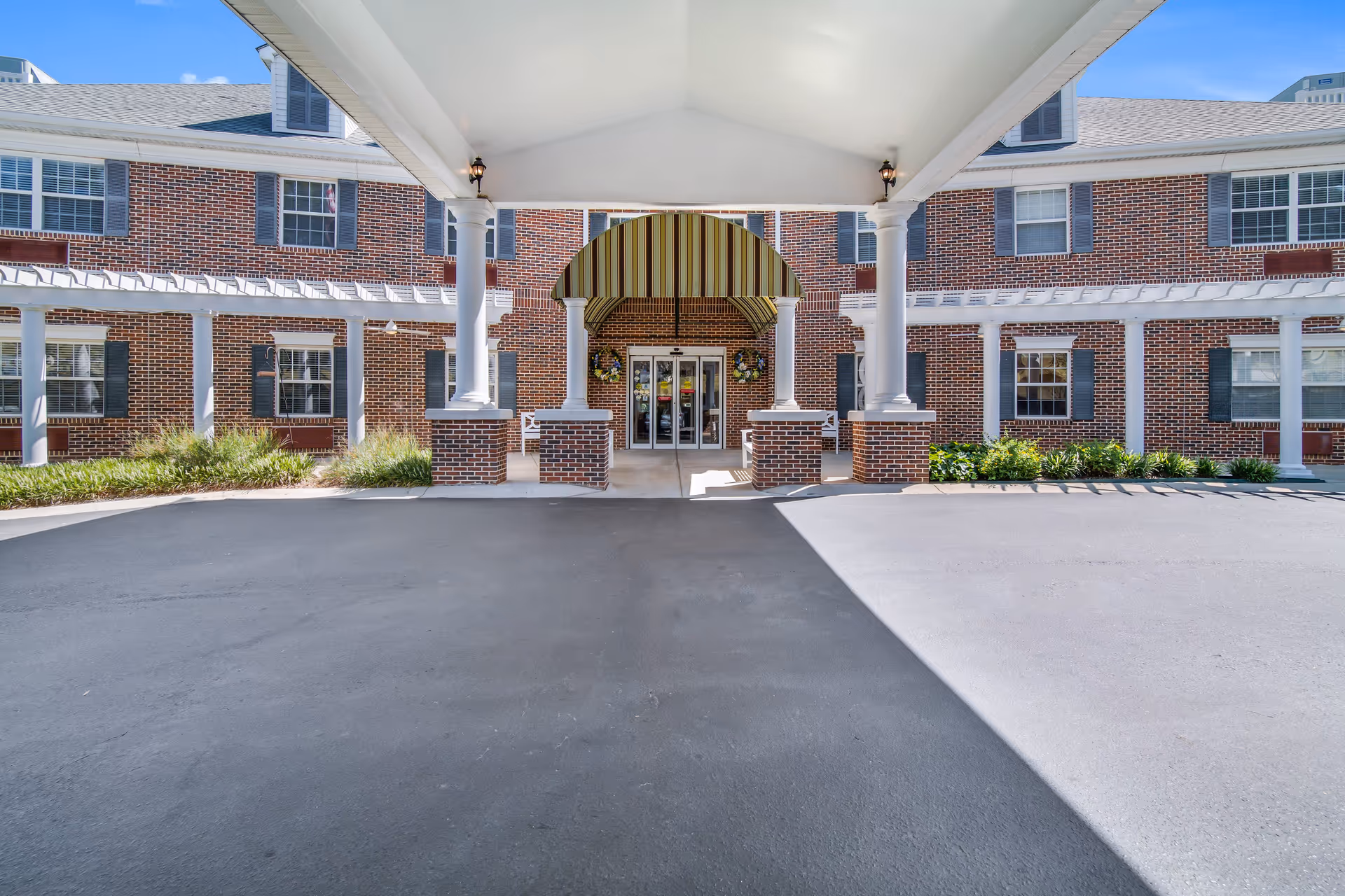 Front entrance of a brick building with white columns supporting a covered driveway. The entrance has double glass doors under a striped awning, with wreath decorations on either side. There are windows with shutters on the building and some landscaping with bushes and plants.
