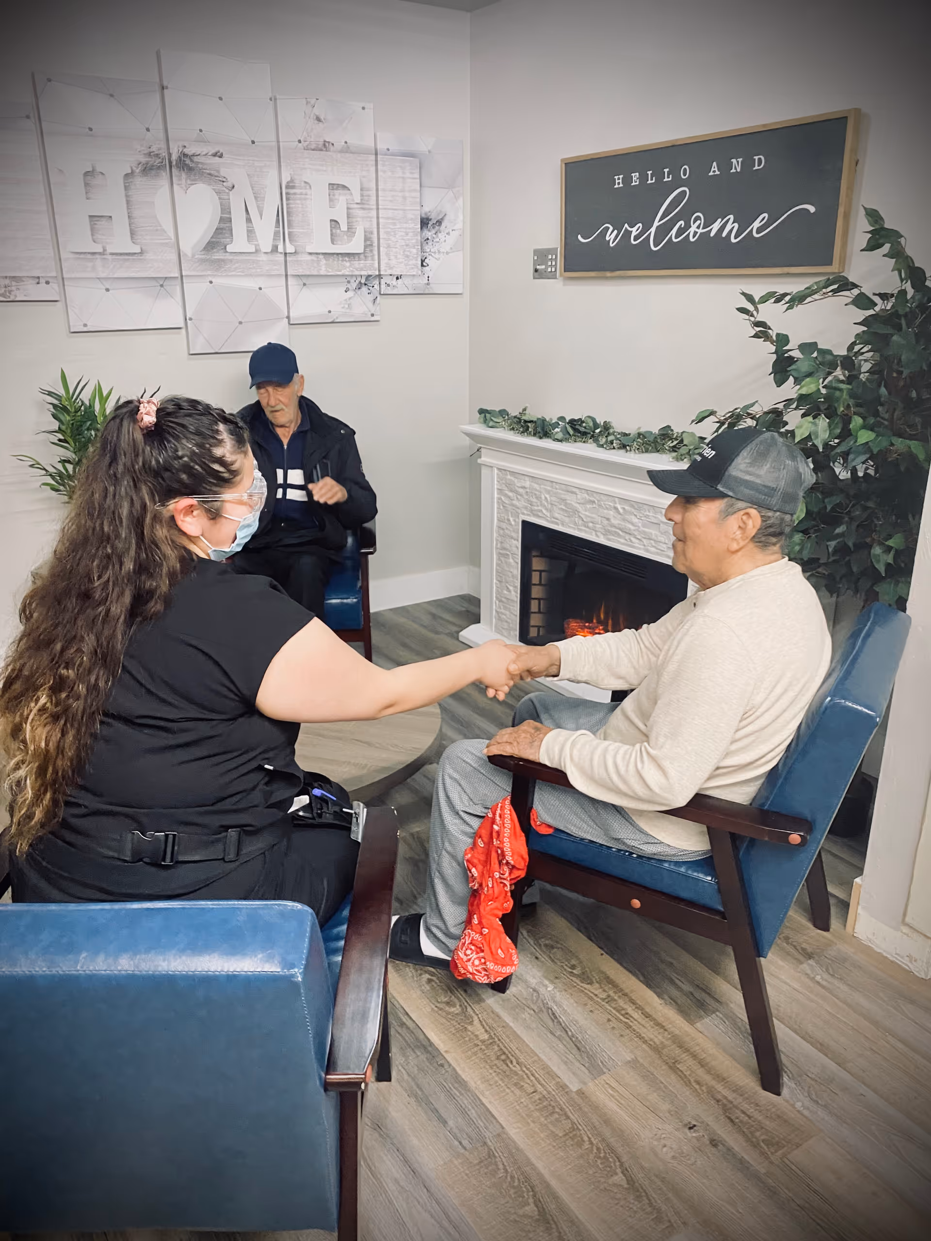 A woman wearing a face mask and protective glasses is sitting in a blue chair and holding hands with an elderly man wearing a cap and light-colored sweater, who is also seated in a blue chair. Another elderly man wearing a dark jacket and cap is sitting in the background. The room has a cozy atmosphere with a white fireplace, a plant, a wall decoration that says 'HOME', and a sign that reads 'HELLO AND welcome'.