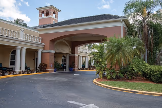 Exterior view of a senior living facility entrance with a covered driveway, columns, and a small tower structure on the roof. The building is surrounded by palm trees and landscaped greenery under a partly cloudy sky.