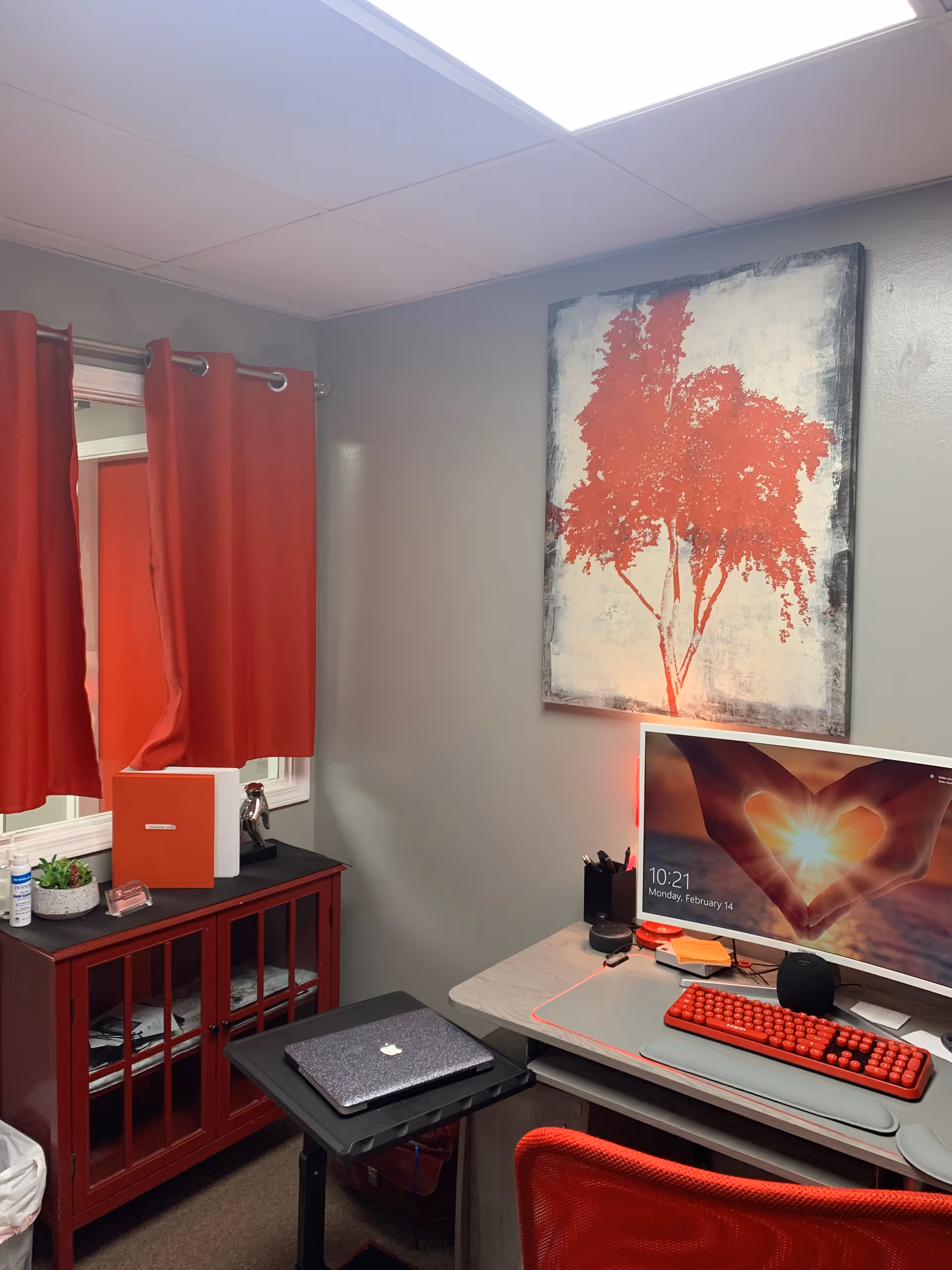 Small interior office space with red curtains, desk, computer, and artwork.
