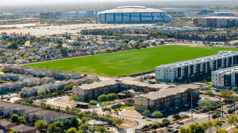 Aerial view of a stadium, surrounding houses, and a central green sports field from above.