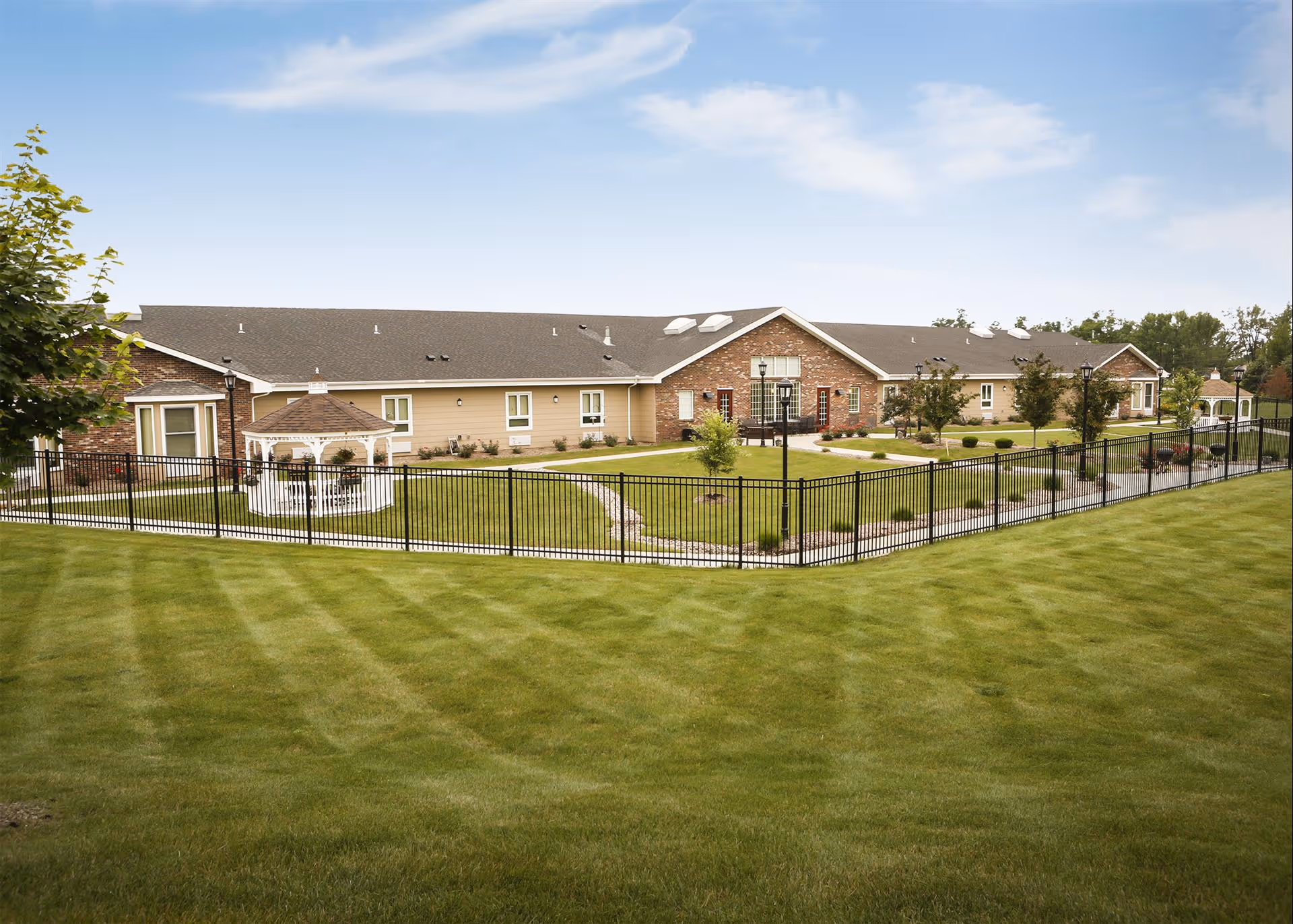 Single-story assisted living building with a manicured lawn, black metal fence, and a white gazebo under a blue sky.