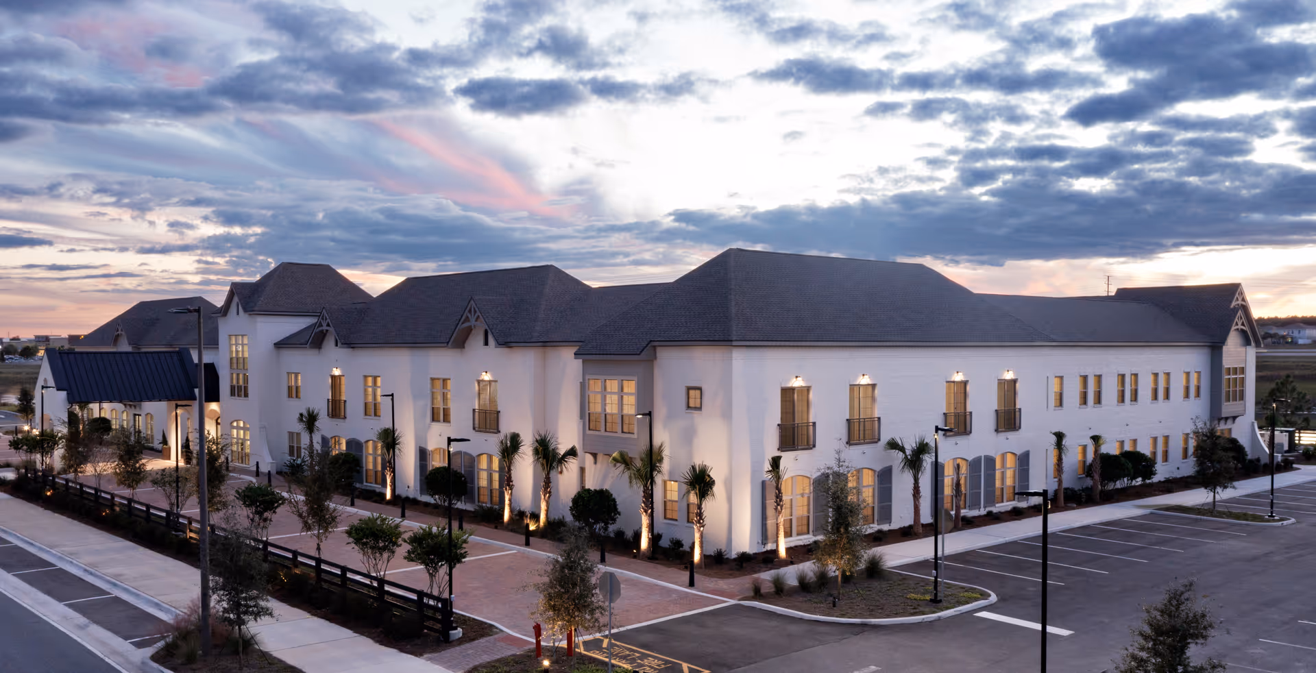 Two-story white senior living facility building photographed at dusk with illuminated windows, palm trees, and an adjacent parking lot.