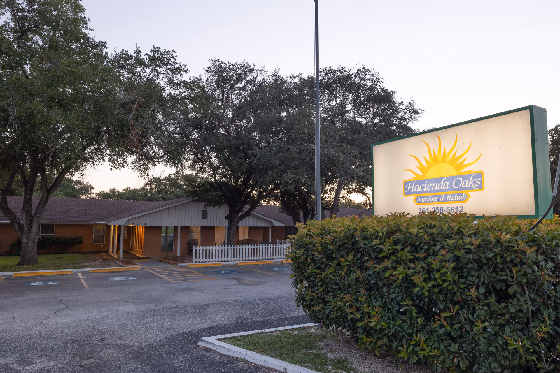 Exterior view of Hacienda Oaks Nursing & Rehabilitation Center during early evening with a lit sign displaying the facility name and phone number, surrounded by trees and bushes, and a parking area with handicap spaces in front.