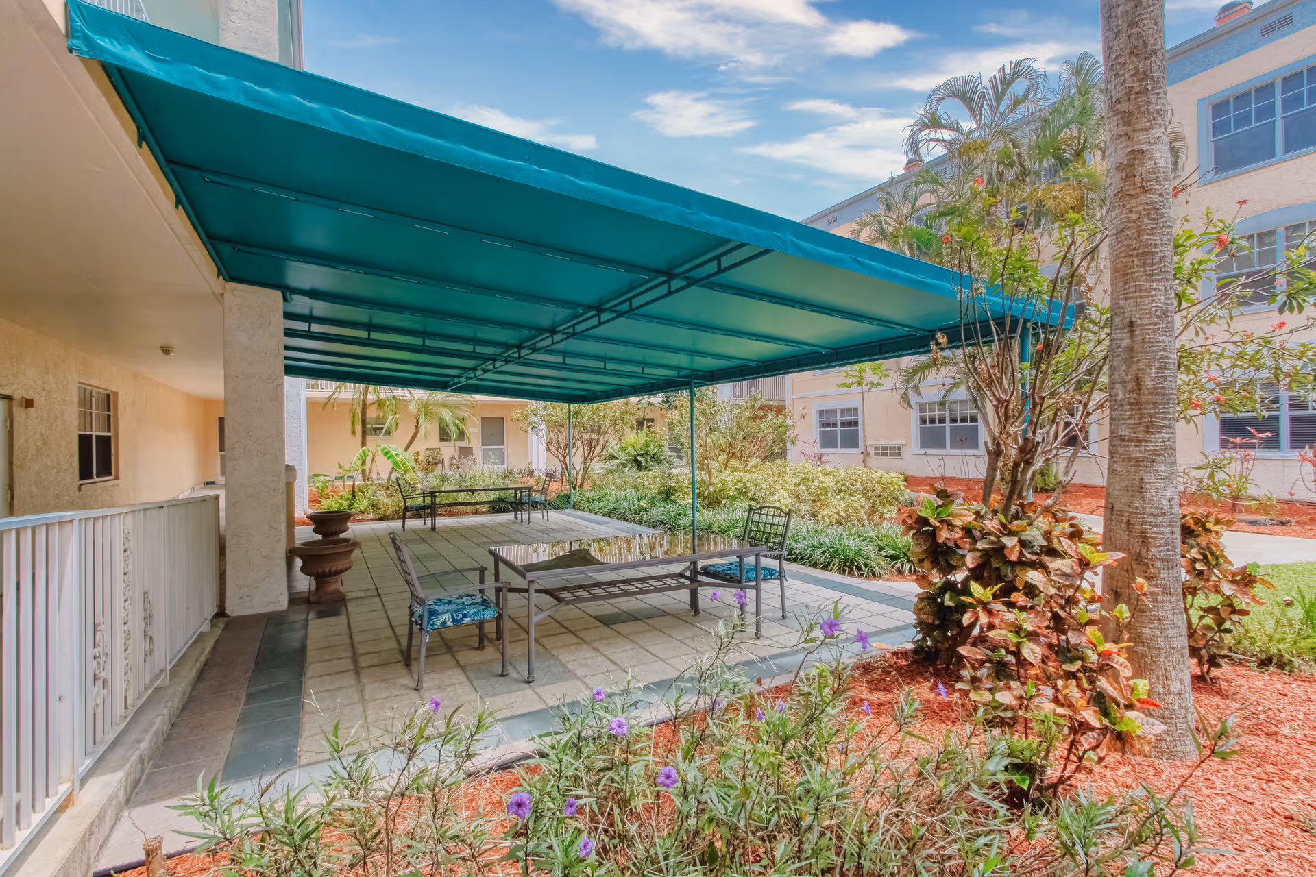 Outdoor patio area at Villa Franciscan with a large green canopy providing shade over metal tables and chairs. Surrounding the patio are landscaped garden beds with various plants and flowers, and the building exterior is visible in the background under a partly cloudy sky.