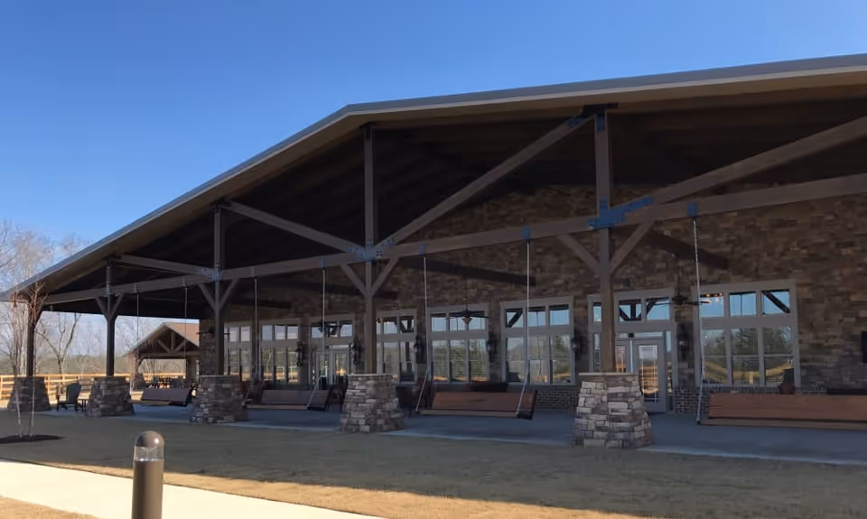 Exterior view of a large building with a covered porch supported by stone pillars and wooden beams. The porch features several wooden porch swings hanging from the ceiling. The building has multiple large windows and doors, and the sky is clear and blue.