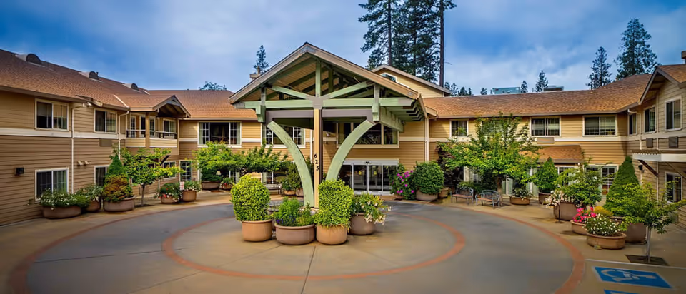 Exterior view of Eskaton Village Grass Valley showing a two-story building with beige siding and a brown roof surrounding a circular driveway with a covered entrance. The area is decorated with various potted plants and trees, and the sky is clear with some tall pine trees in the background.