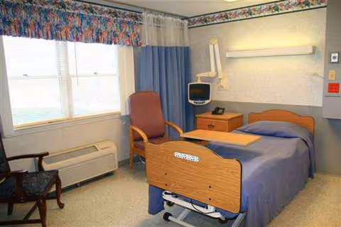 Single patient bedroom in a care facility with blue bed, chair, and medical equipment.