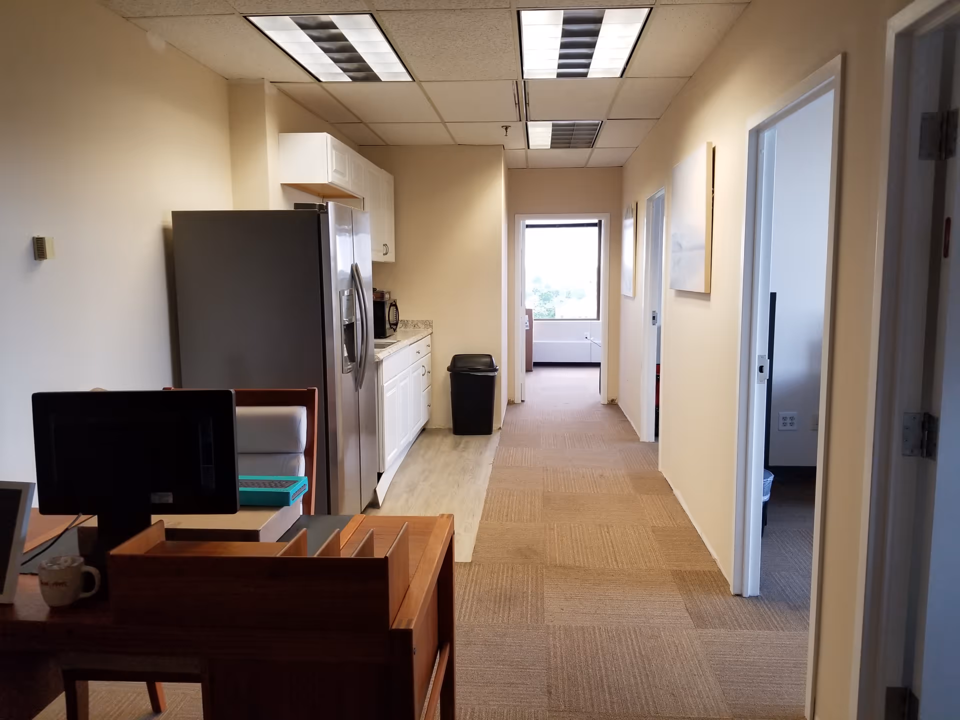 Narrow hallway with kitchenette, refrigerator, microwave, and fluorescent ceiling lights in a senior care facility.