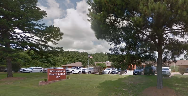 Front lawn and parking area of Whispering Pines Lodge with a sign, American flag, parked cars, and surrounding trees.