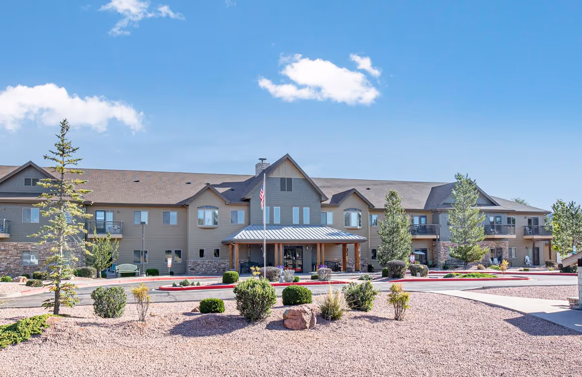 Exterior view of Majestic Rim Retirement Living facility showing a two-story building with a covered entrance, surrounded by landscaped grounds with bushes, trees, and gravel. The sky is clear with a few clouds.