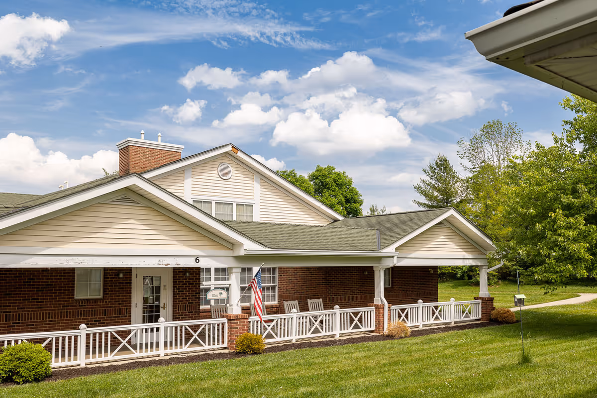 Exterior view of a single-story brick building with beige siding and a green roof under a partly cloudy blue sky. The building has a covered porch with white railings, several rocking chairs, an American flag, and a sign that reads 'Elm'. The surrounding area includes green grass, bushes, and trees.