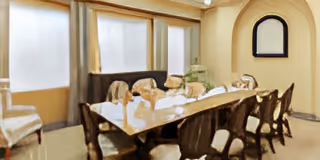 A dining room with a long rectangular table surrounded by multiple chairs. The table is set with plates and glassware. There are large windows with curtains on one side and a mirror on the wall.