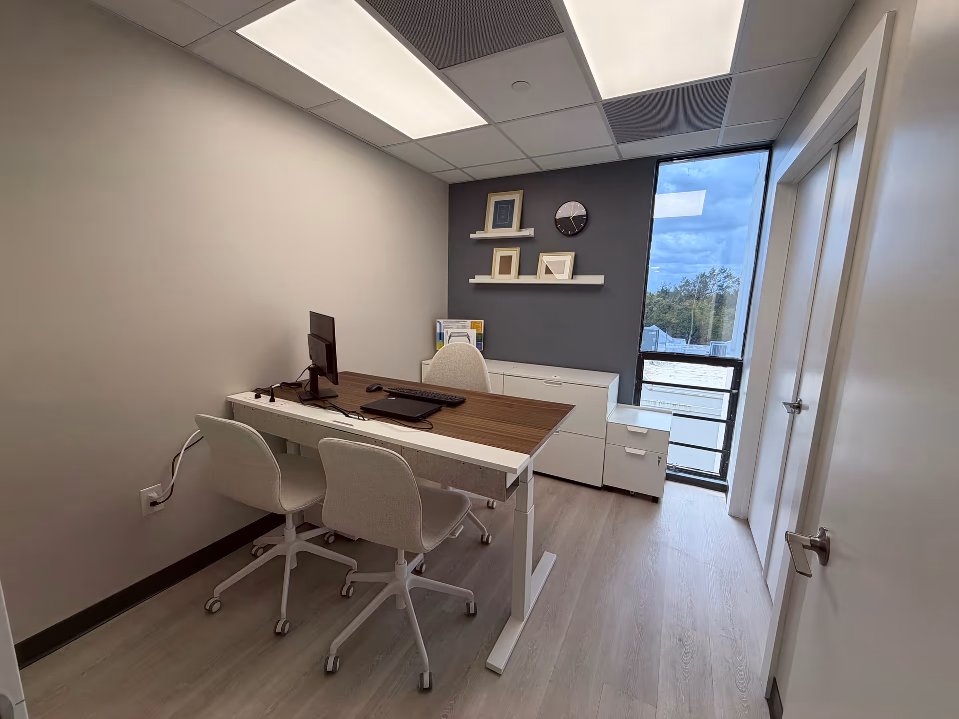 A small office with a desk, computer, white chairs, and wall shelves near a window.