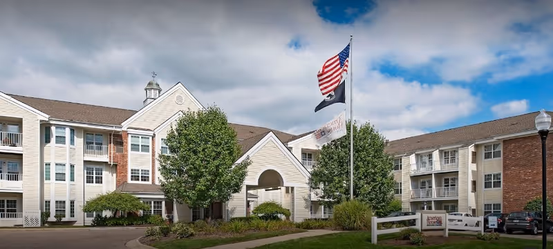 Exterior view of Baldwin House Senior Living Hazel Park, showing entrance, flags, and trees.