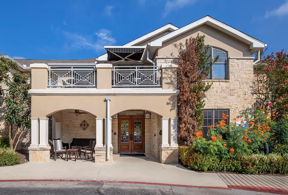 Front exterior of a two-story beige stone building with a covered entrance, balcony, seating area, and landscaped flowers.