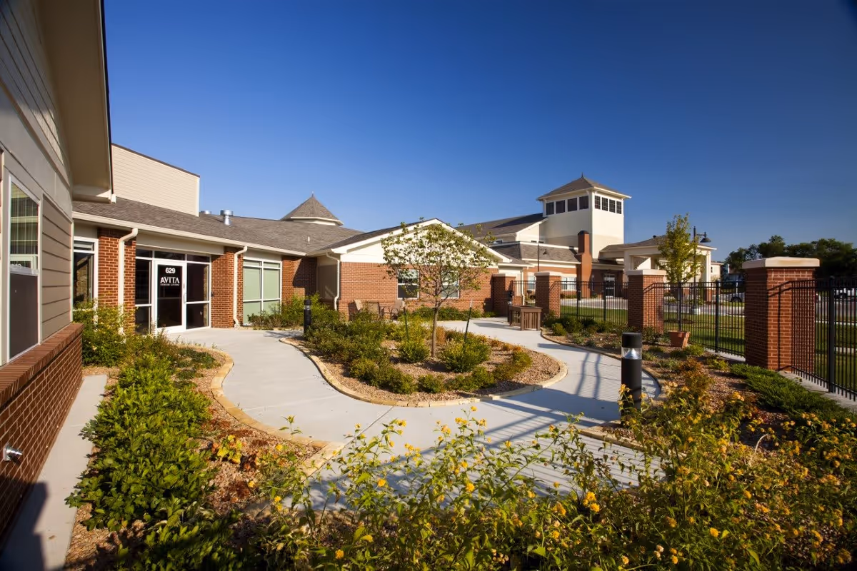 Courtyard and entrance of Avita Senior Living with winding walkways, landscaped beds, and a brick building under a clear blue sky.