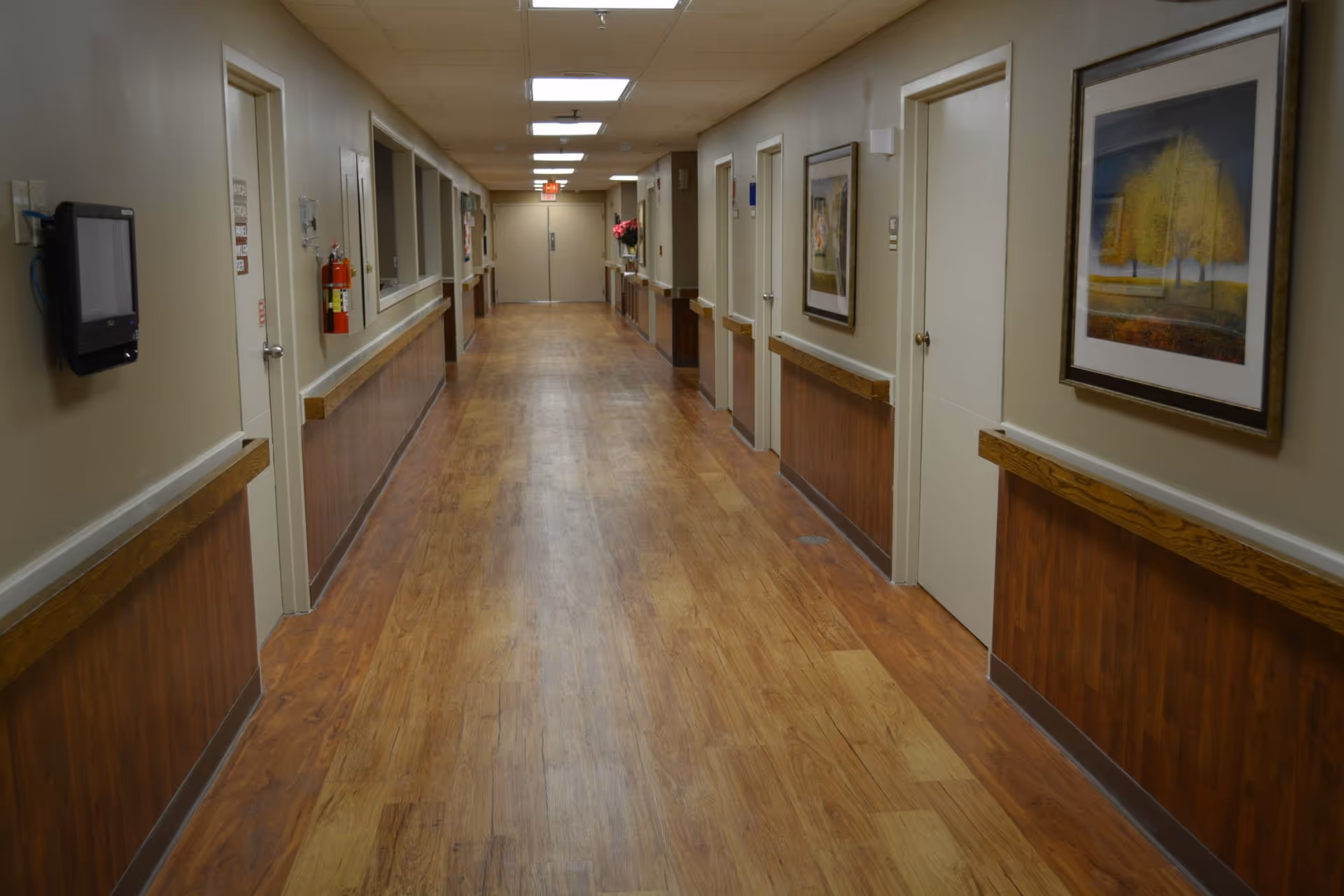 A long, empty hallway in a nursing center with wooden flooring and beige walls. The hallway has several closed doors on both sides, handrails along the walls, framed artwork, a fire extinguisher, and ceiling lights. Double doors are visible at the far end of the corridor.