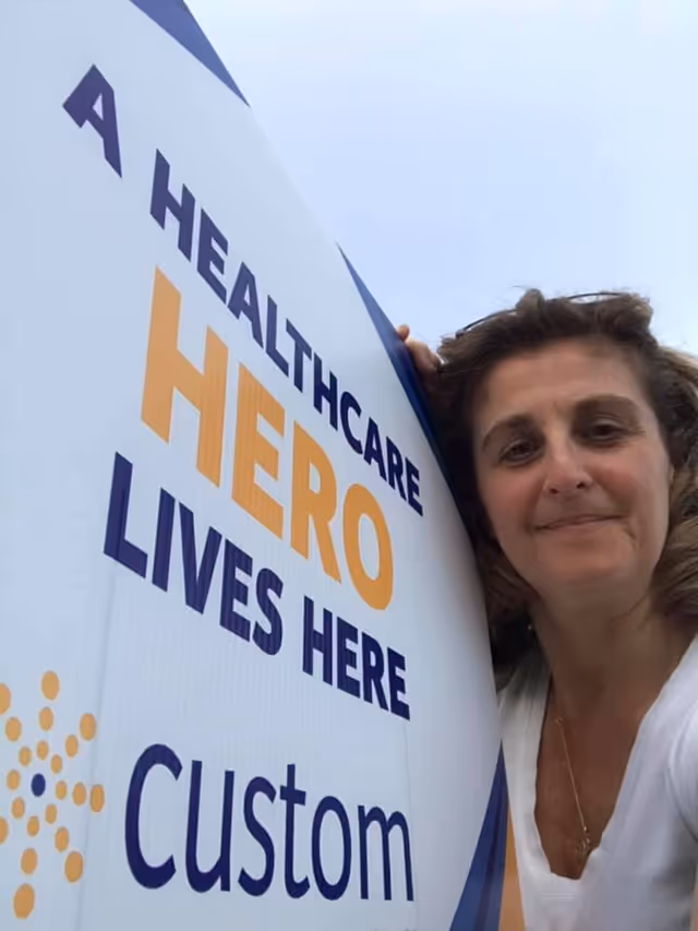 Smiling woman beside a large Custom Hospice Healthcare Hero sign outdoors.