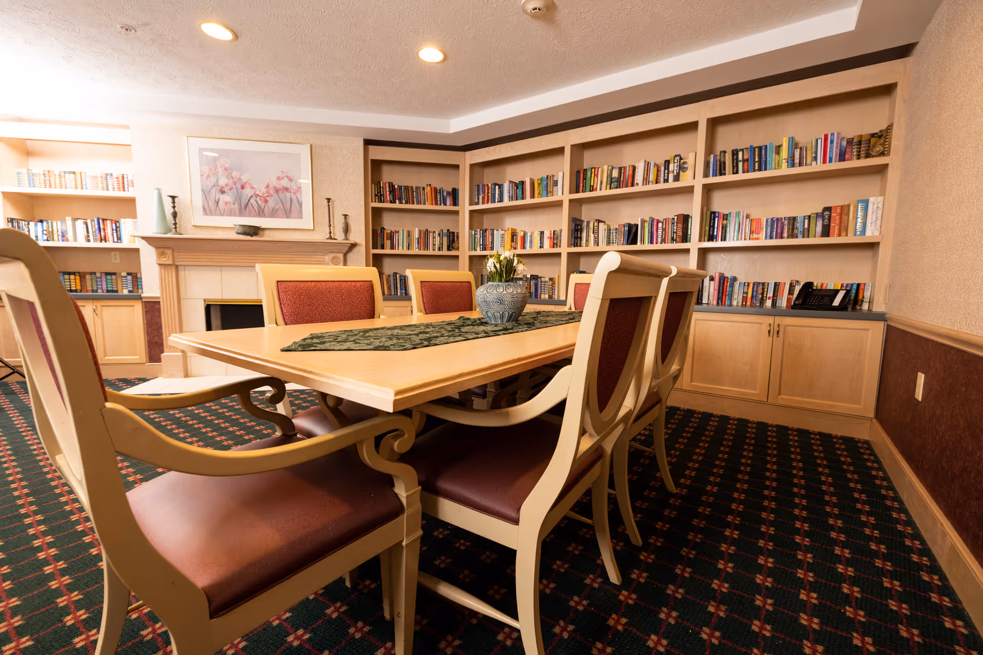 A cozy room with a wooden table surrounded by six chairs with red cushions. The room features built-in bookshelves filled with books, a fireplace with a floral painting above it, and a patterned carpet. A decorative vase with flowers is placed on the table.