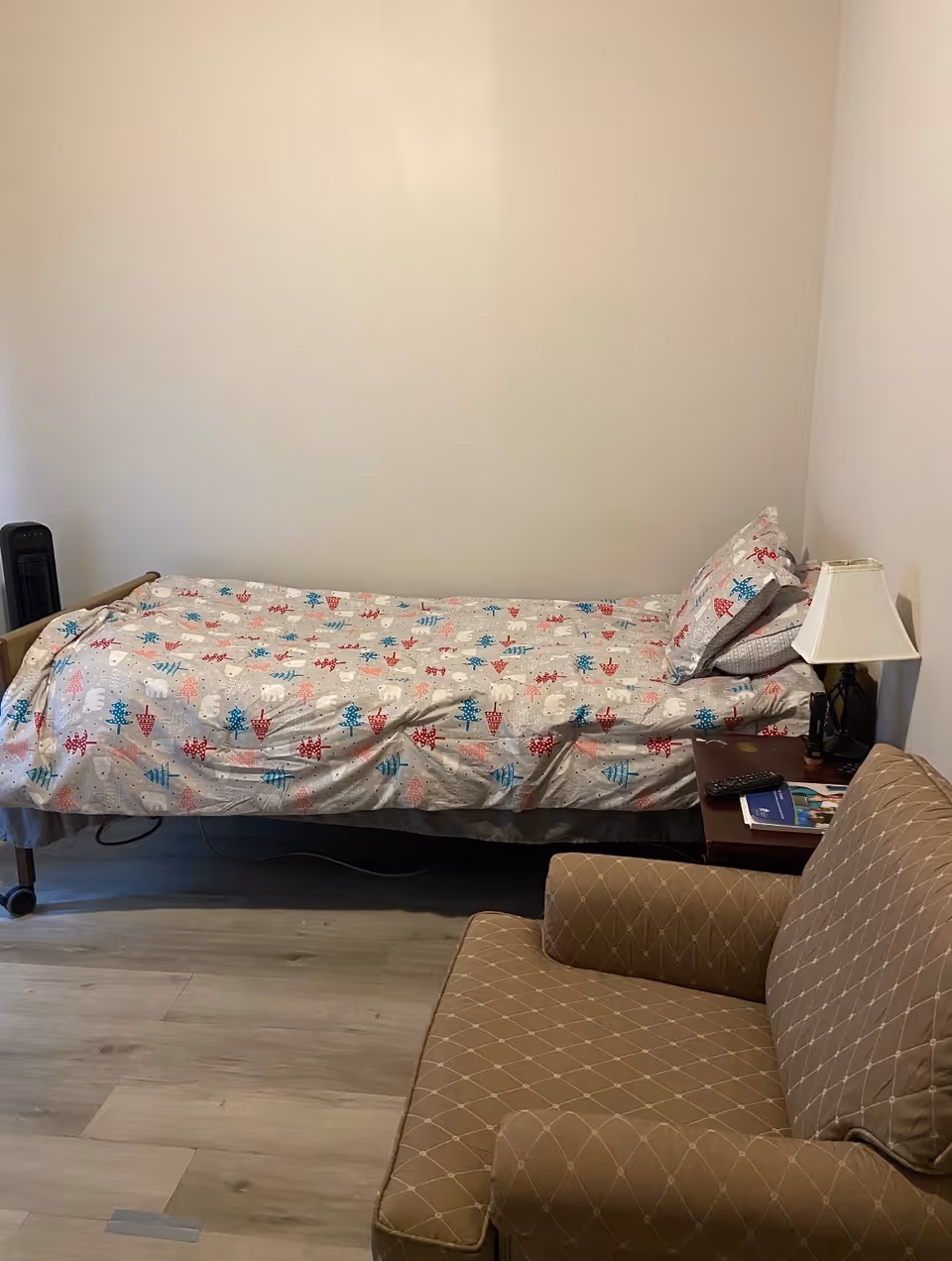 A single bed with patterned linens, bedside table, lamp, and a brown chair in a bedroom.