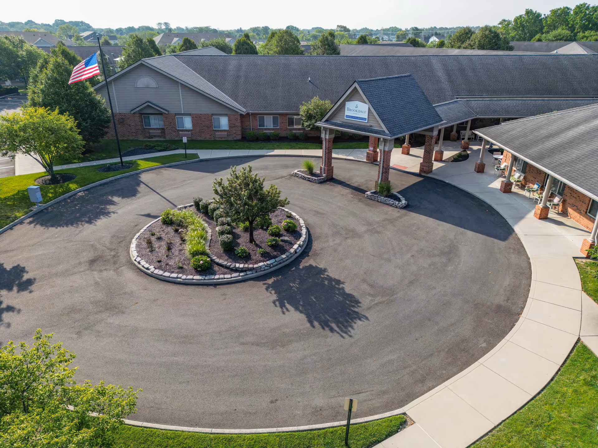 Aerial view of the entrance to Brookdale Pinnacle senior living facility showing a circular driveway with a landscaped island in the center, an American flag on a flagpole, and a covered drop-off area supported by brick pillars. The building is a single-story structure with a brick and siding exterior surrounded by trees and greenery.