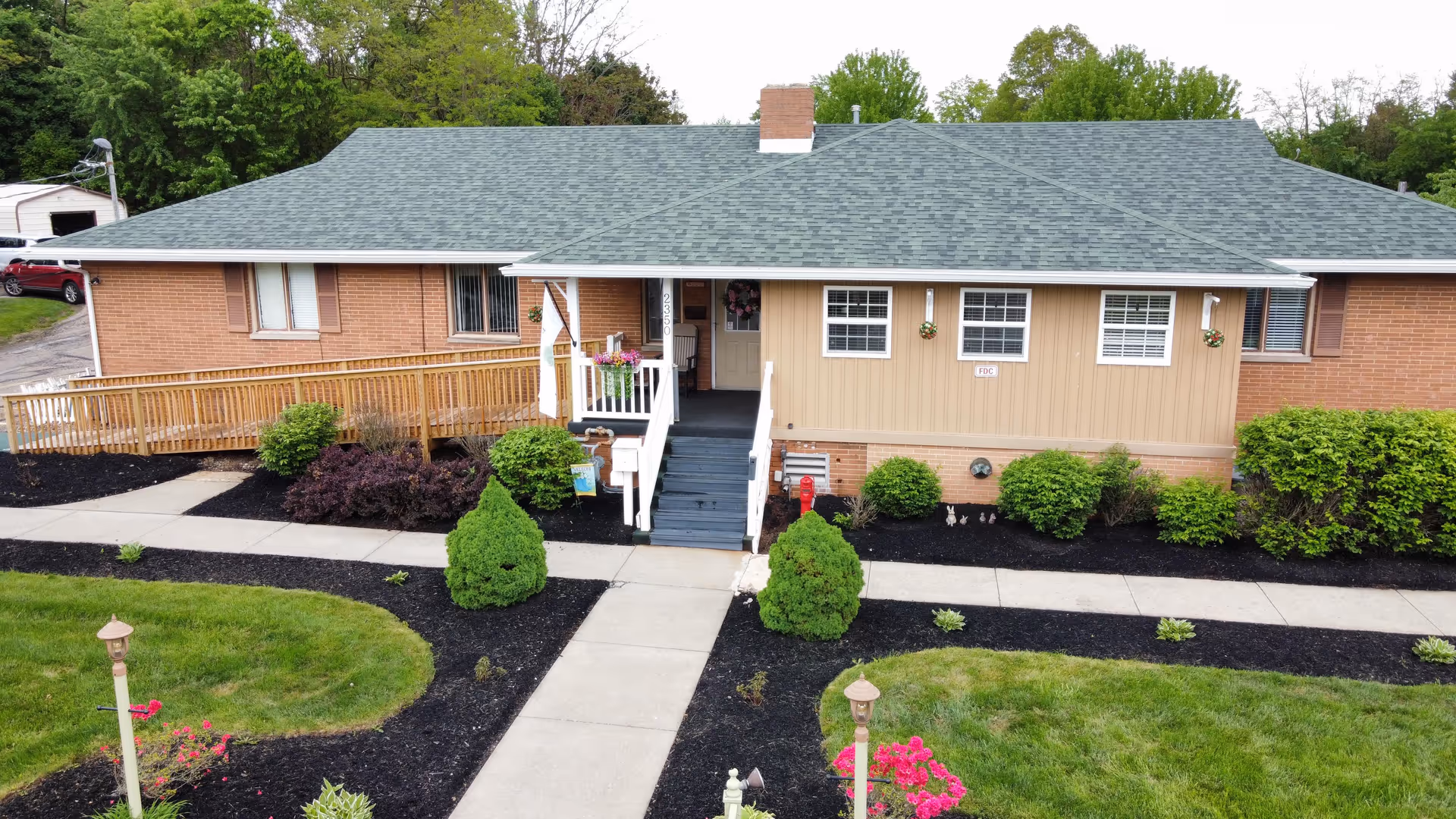 Front exterior view of a single-story senior living community building with a green shingled roof, brick and beige siding, a small porch with stairs and a wheelchair ramp, surrounded by landscaped bushes, flower beds, and a concrete walkway.