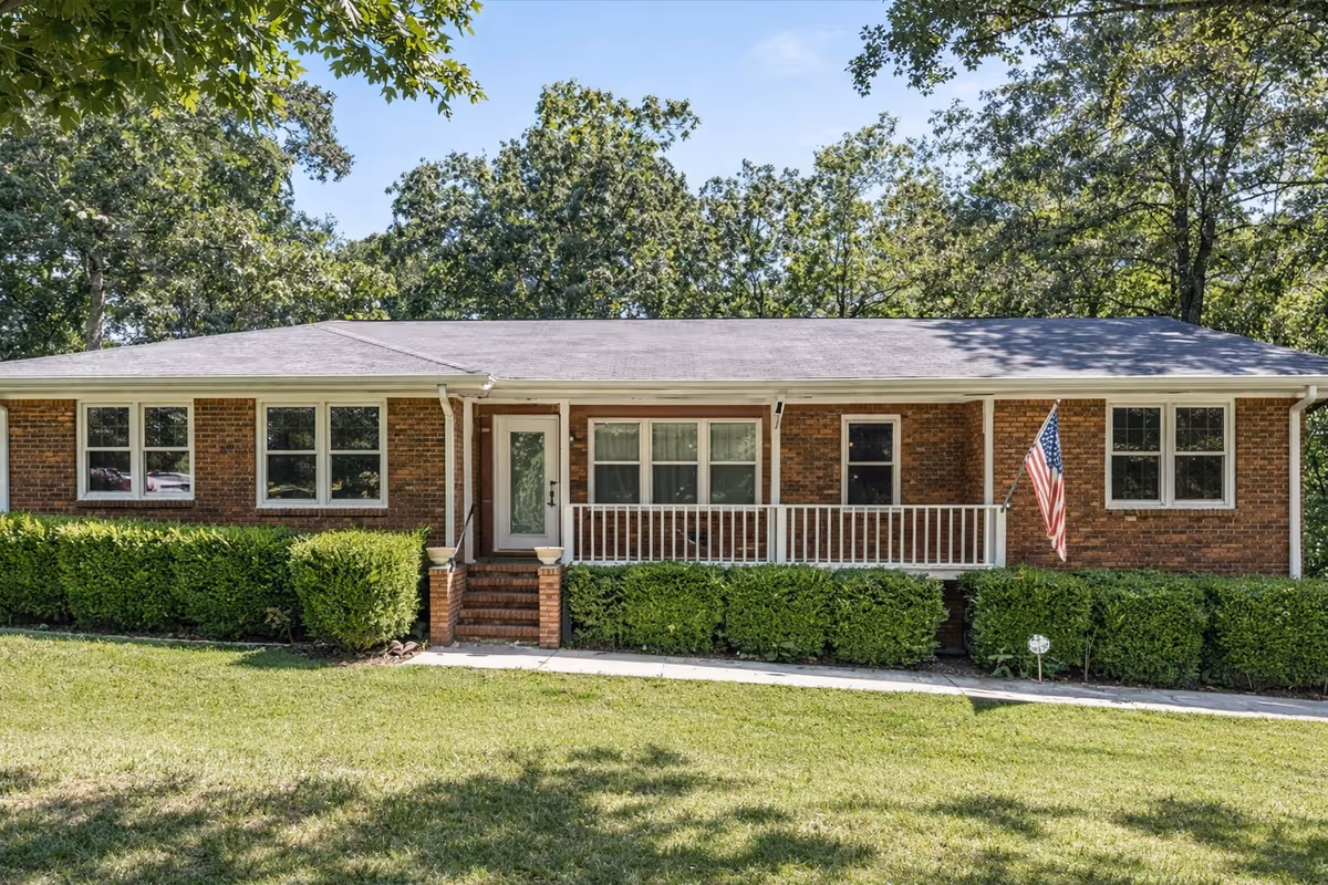 Single-story brick senior living facility with a front porch, hedges, and an American flag.