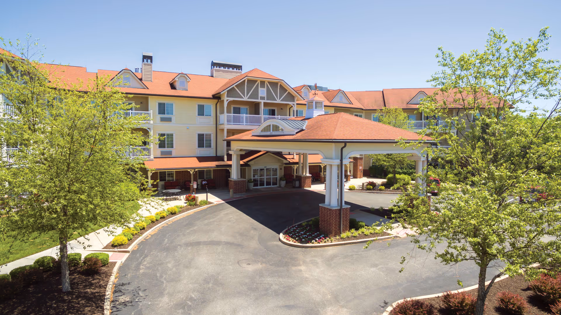 Exterior view of a senior living facility with a large covered entrance, surrounded by trees and landscaping under a clear blue sky.