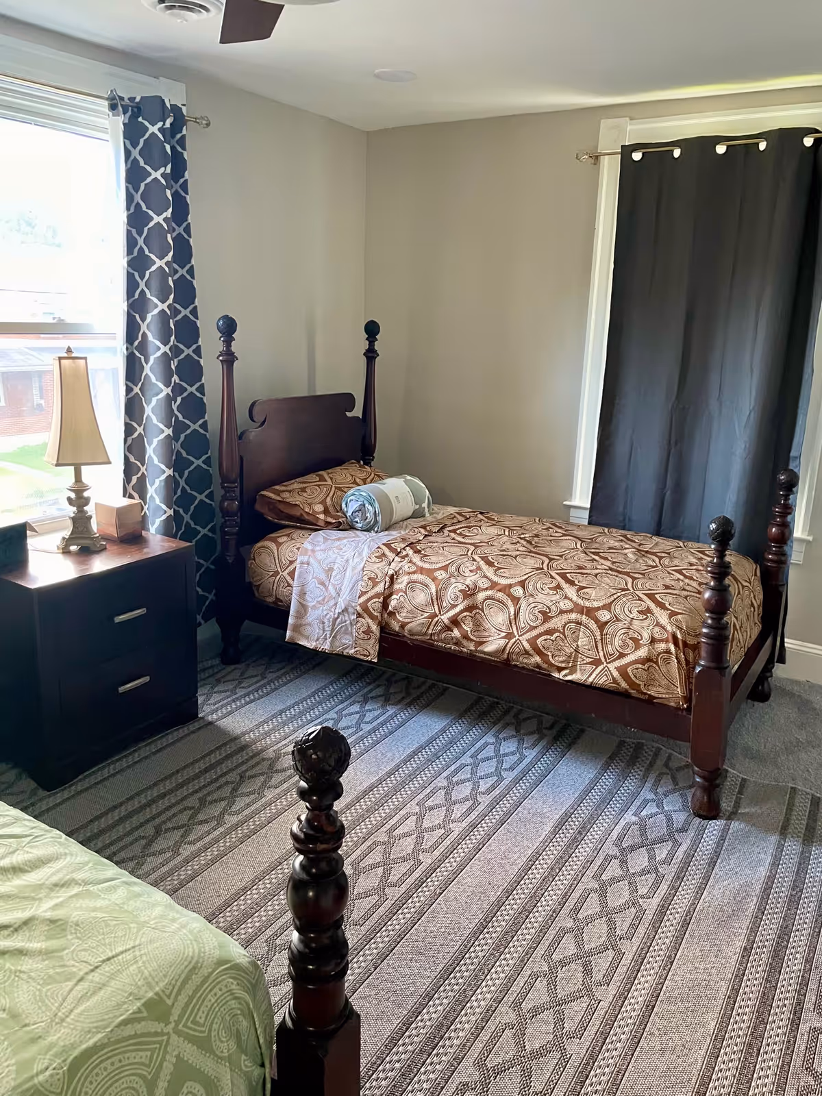 A bedroom with a wooden four-poster bed, nightstand, lamp, and patterned curtains.