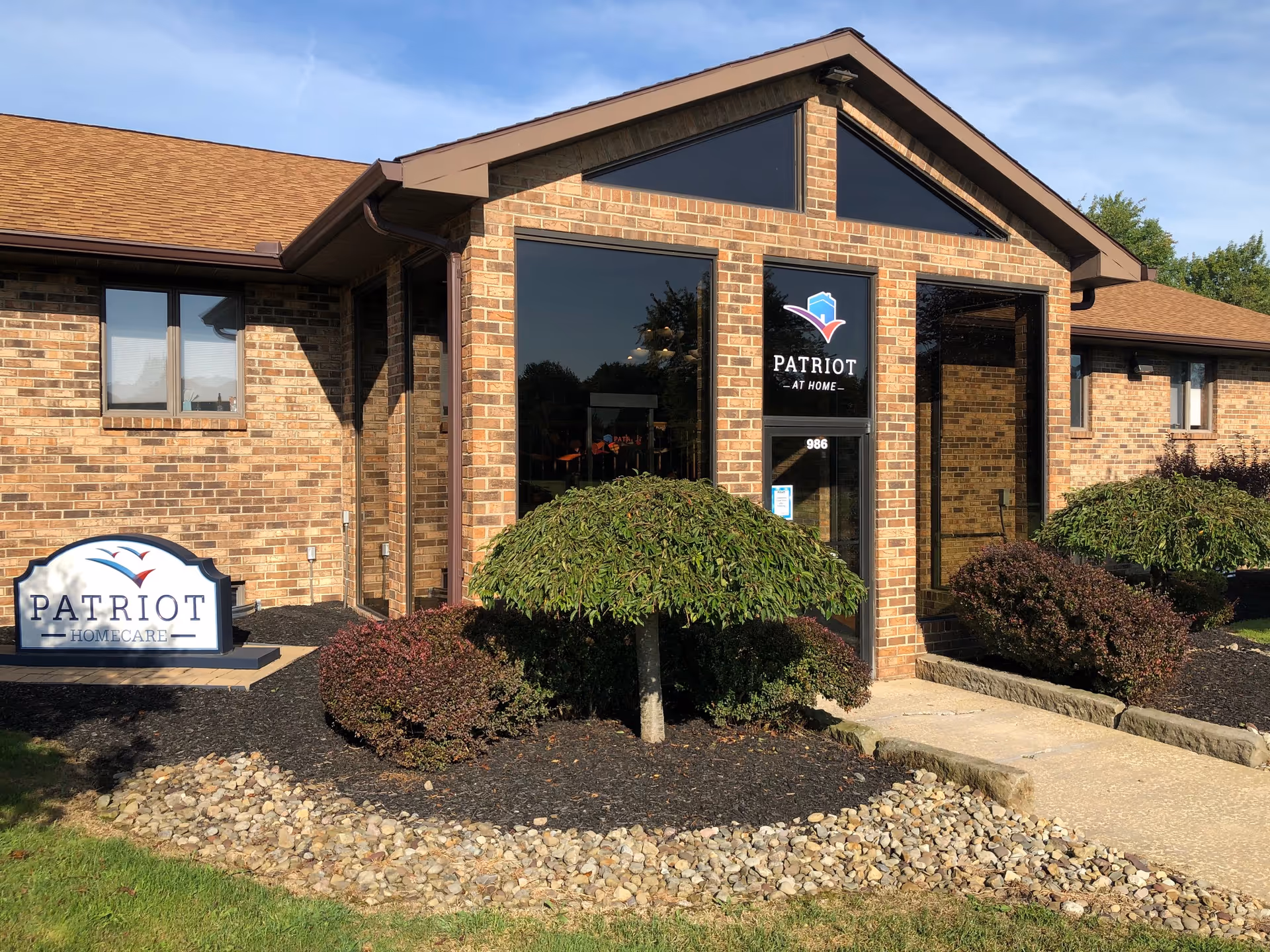 Brick Patriot at Home facility exterior with glass entrance and manicured shrubs.