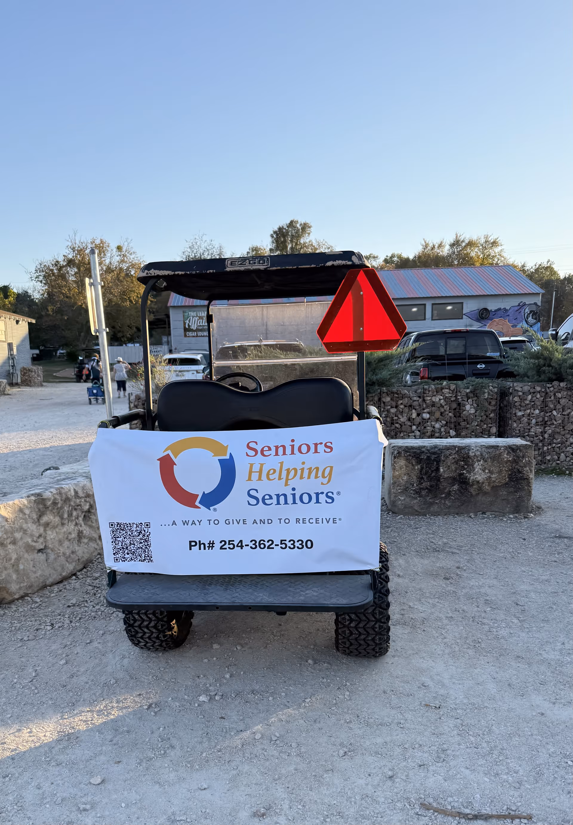 Golf-cart with a Seniors Helping Seniors banner parked on a gravel outdoor lot.
