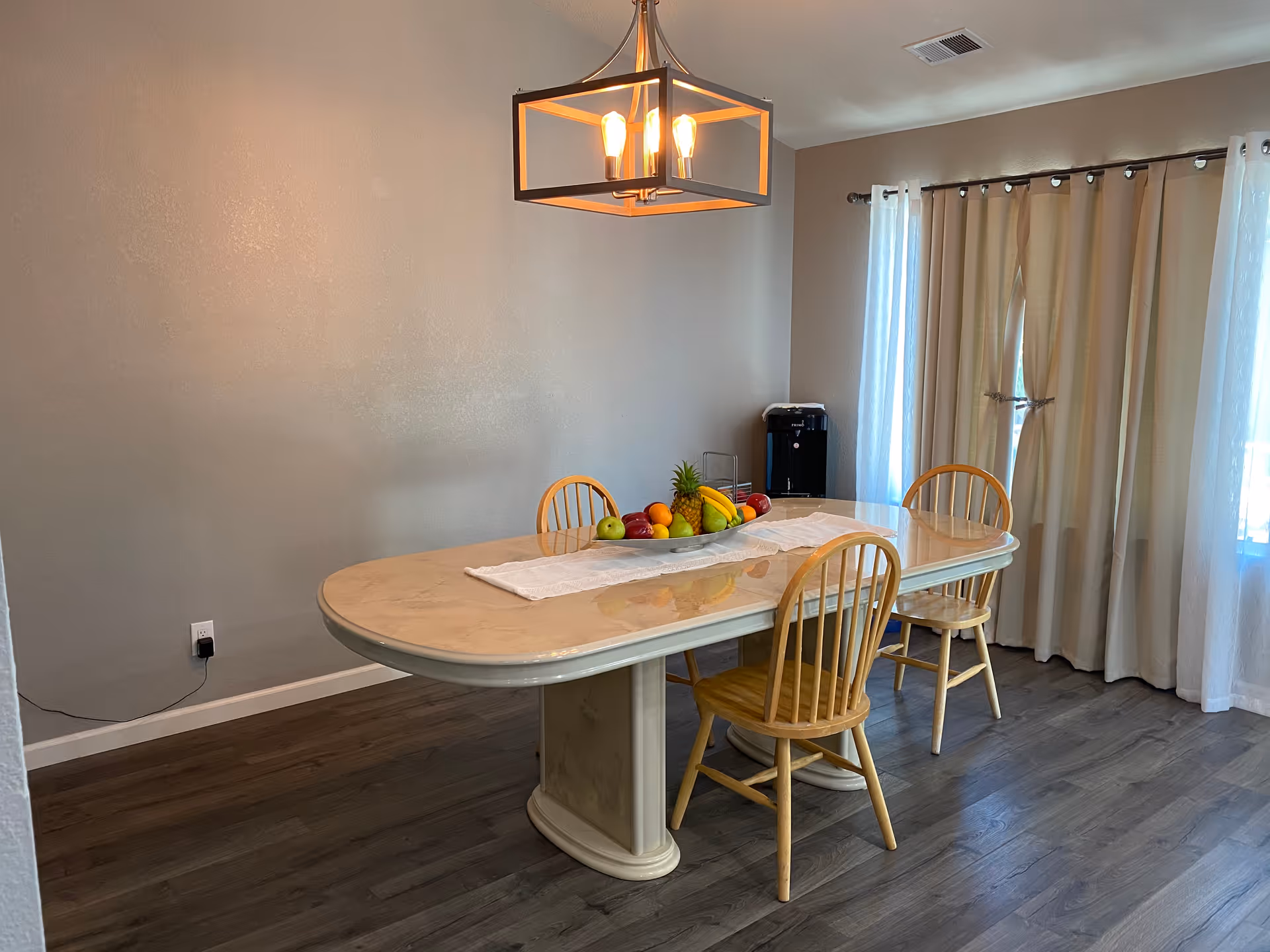 Dining room with an oval table, wooden chairs, a fruit centerpiece, pendant light, and curtained windows.