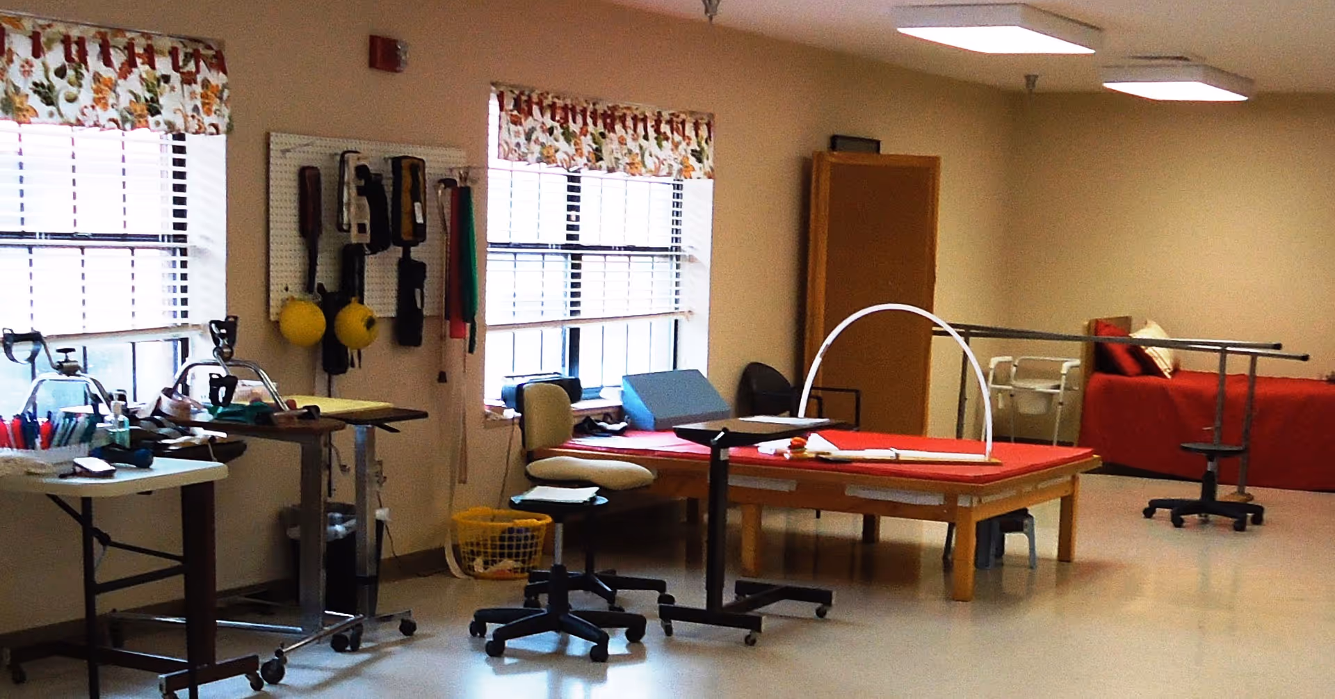 Rehabilitation therapy room with treatment tables, exercise equipment, chairs, and windows.
