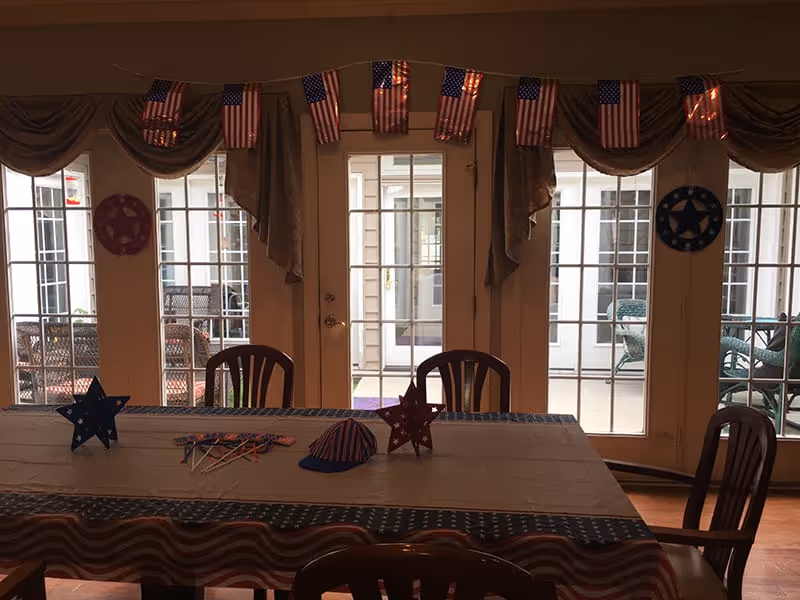 A dining room decorated with American flag-themed items including a tablecloth, small flags hanging above the windows, and star-shaped decorations on the table. The room has large glass doors and windows looking out onto a patio area with outdoor furniture.