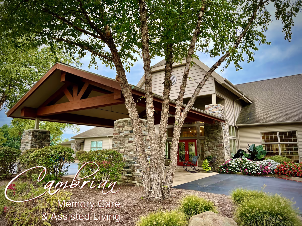 Exterior view of Cambrian Assisted Living and Memory Care Tecumseh building entrance with a covered driveway, stone pillars, a tree in the foreground, and landscaped flowers and bushes around the entrance.