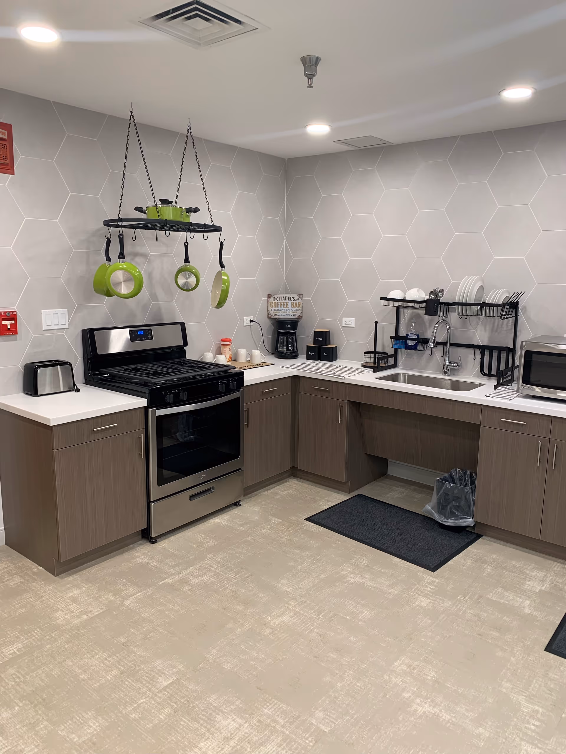 Modern kitchen area with a stainless steel stove and oven, a hanging rack with green pots and pans, a coffee maker on the countertop, a toaster, a microwave, and a sink with dishes drying on a rack. The walls have light gray hexagonal tiles and the floor is covered with beige patterned flooring.
