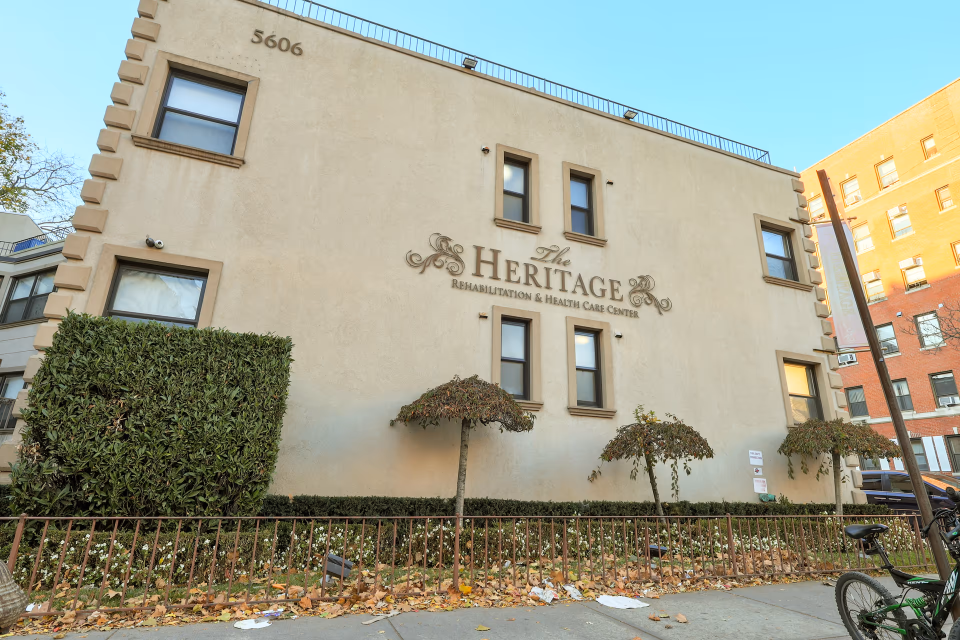 Exterior front of The Heritage Rehabilitation & Health Care Center building with signage, trimmed trees and a sidewalk.