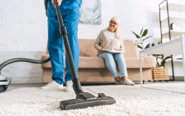 Vacuum cleaner in the foreground; elderly woman sits on a couch in the living area.