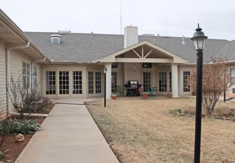 A paved walkway through a grassy courtyard leads to the covered entrance of a single-story senior living building with lamp posts, patio furniture, and French doors.