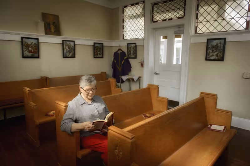 An elderly person sits in wooden church pews reading a book in a chapel-like room.