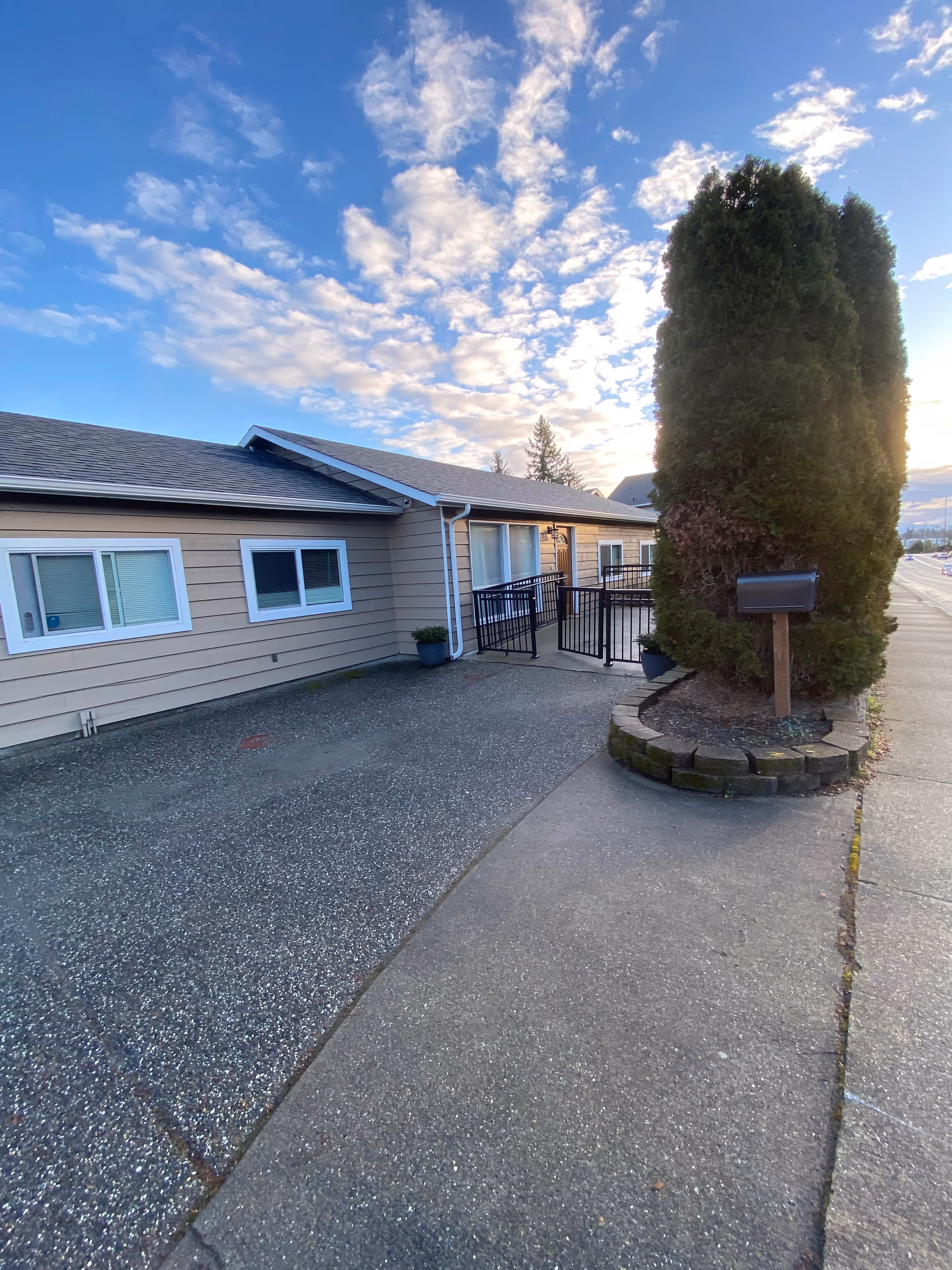 Single-story beige facility exterior with windows, railing, and a tall evergreen tree.