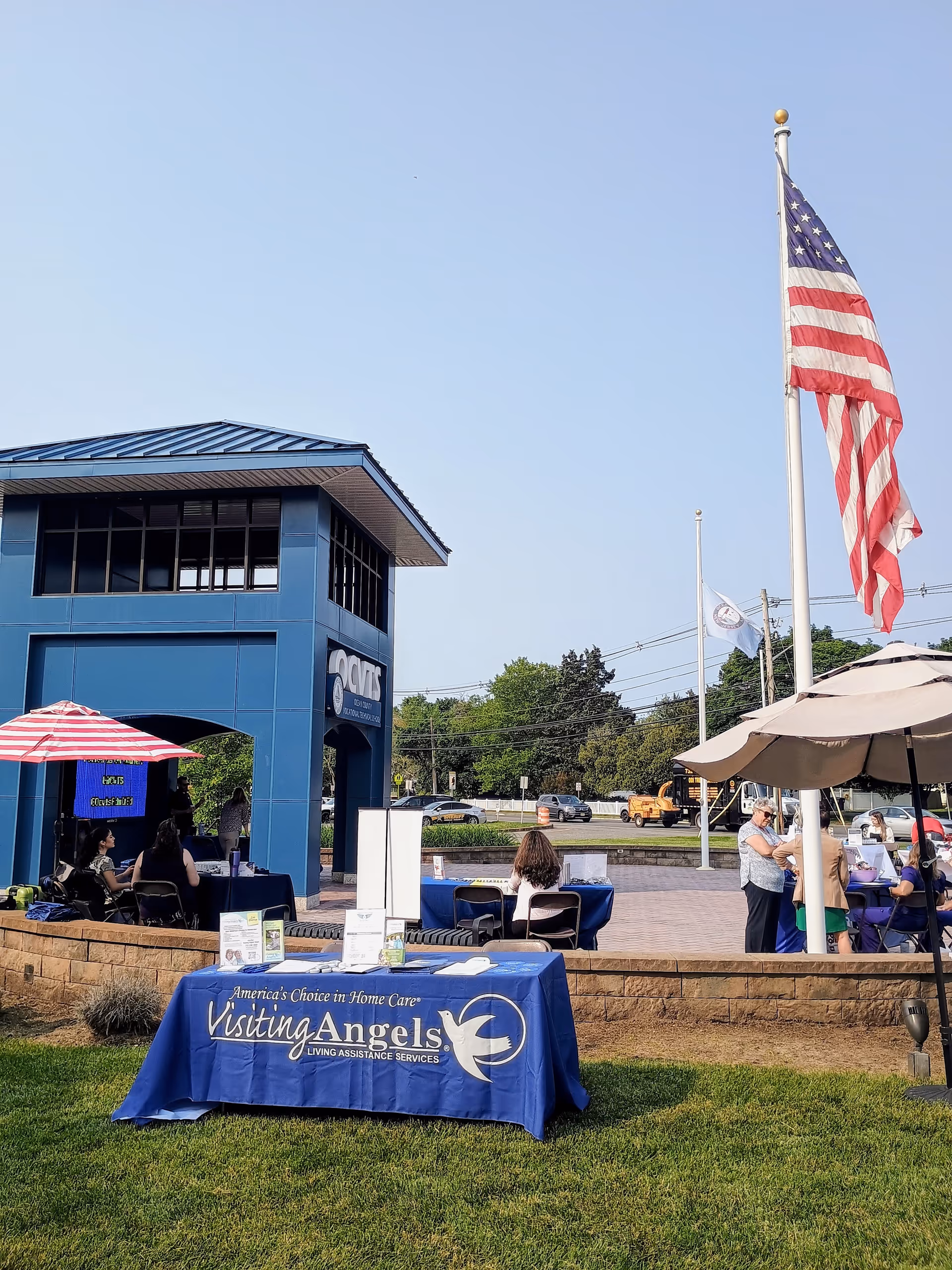 Outdoor event at Visiting Angels facility with blue tables, umbrellas, and an American flag.