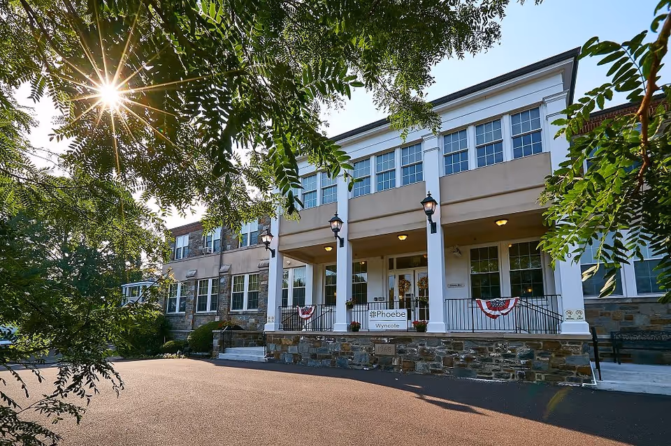 Sunlit front entrance of the Phoebe Wyncote building with a columned portico, stone facade, and decorative bunting.