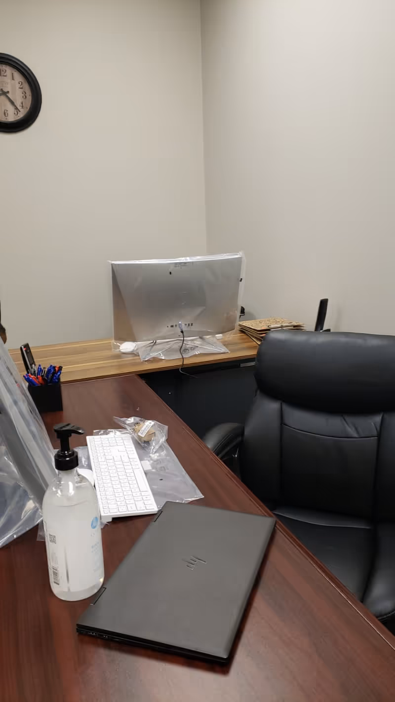 Interior office desk with computer monitor, keyboard, laptop, sanitizer, and a black leather chair.