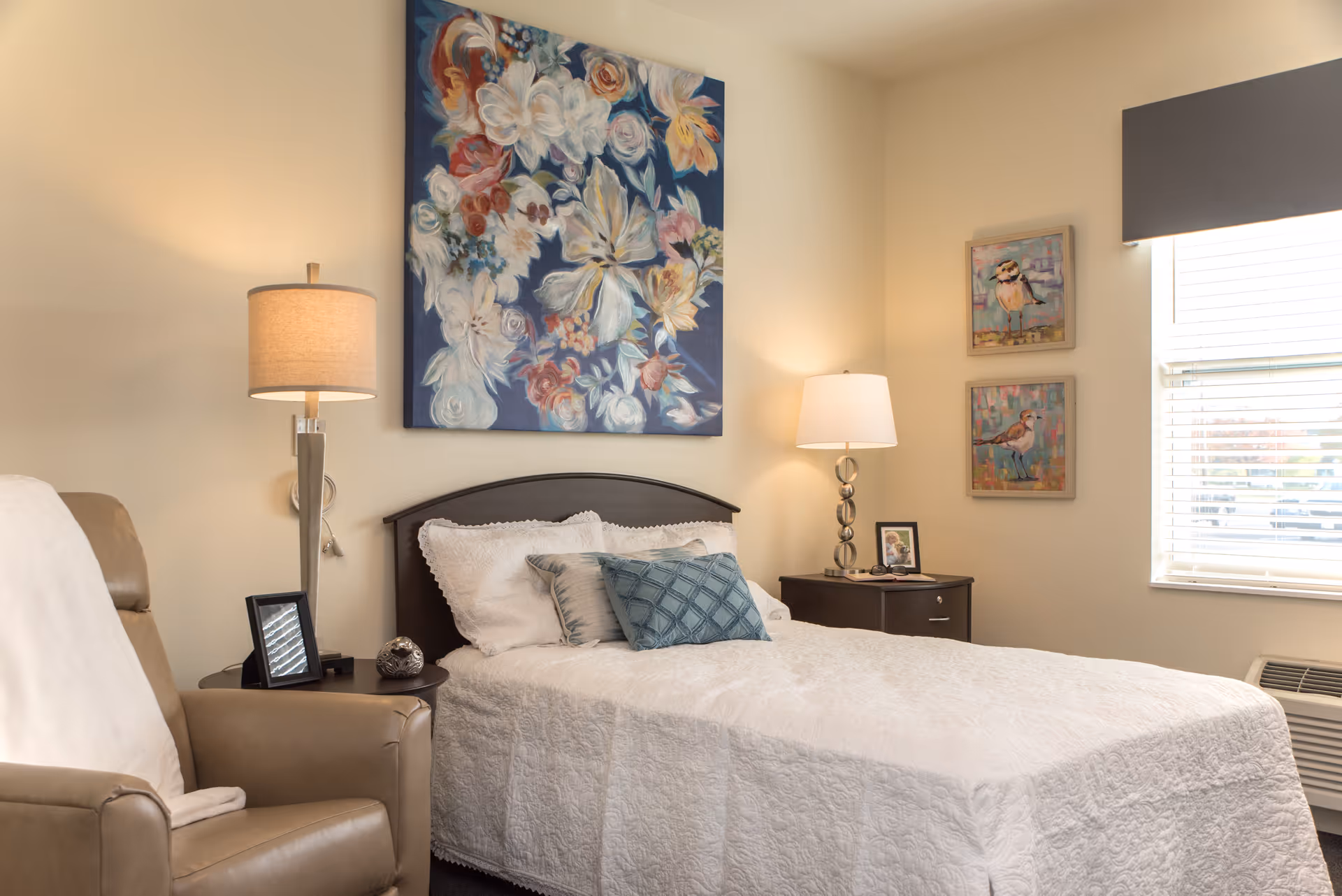 Sunlit bedroom with a made bed, patterned pillows, bedside tables with lamps, an armchair, and floral artwork above the headboard.