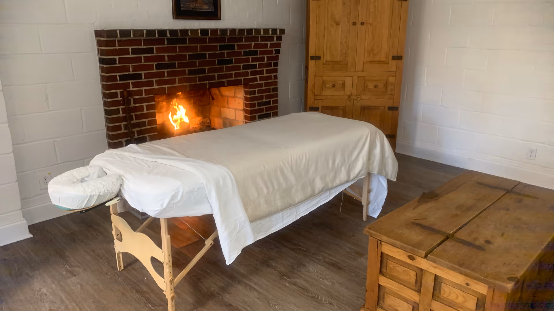 Massage therapy room in a senior living facility with a fireplace, massage table, and wooden cabinet.