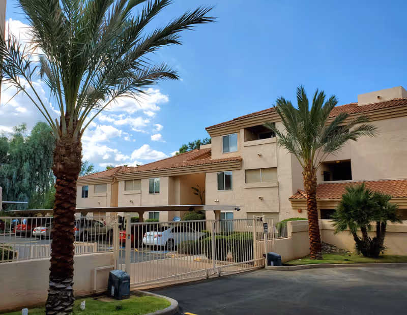 Exterior view of a Jerusalem Assisted Living Facility with a gate, palm trees, and parked cars.