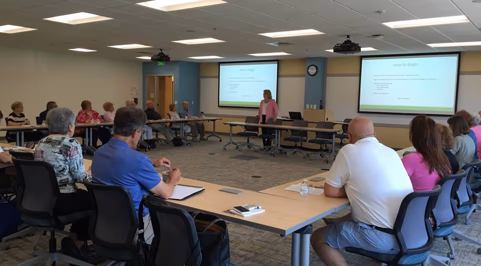 A group of elderly people seated around a large U-shaped conference table in a well-lit meeting room, attentively listening to a woman standing and presenting in front of two projection screens displaying a presentation titled 'How to Begin'.