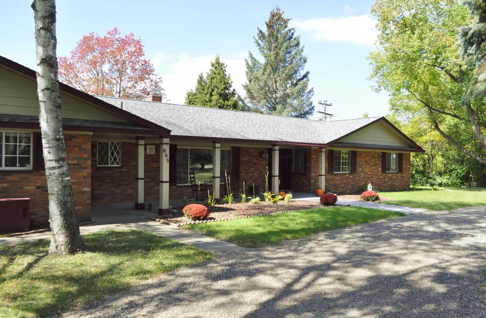 Single story brick senior living facility exterior with a porch, lawn, and autumn landscaping.